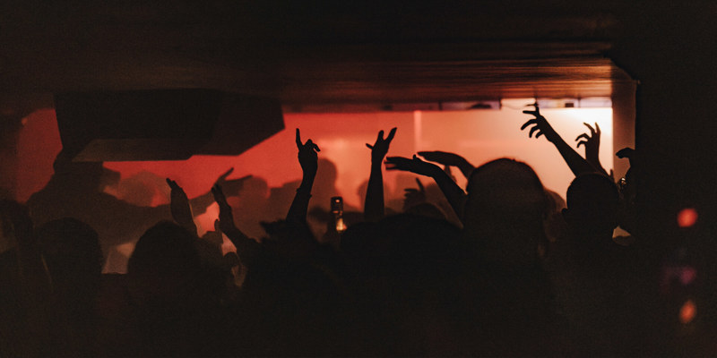 Crowd dancing in a dimly lit basement venue with raised hands silhouetted against warm red and orange background lighting.
