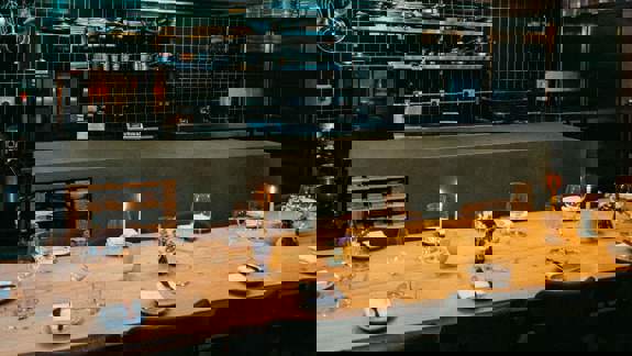 A long wooden dining table set with candles, glasses, and white plates. Behind it is a kitchen with a stainless steel counter and tiled walls.