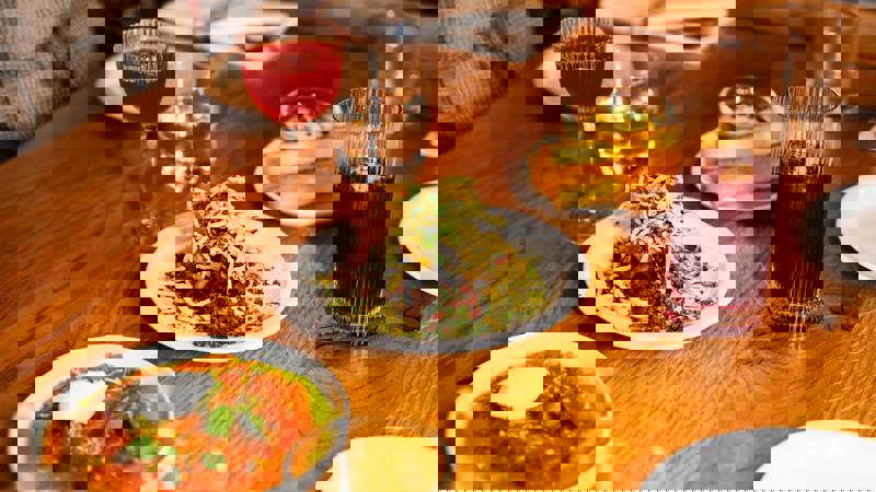 A close-up of a wooden table laden with various plates of food and drinks. The dishes include a salad with shredded vegetables, a bright orange dish, and polenta fries, with wine glasses and tumblers.