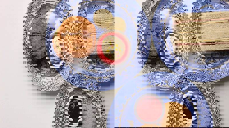 Plates of sandwiches, scones with cream and jam, and assorted desserts on patterned plates on an off-white table setting.