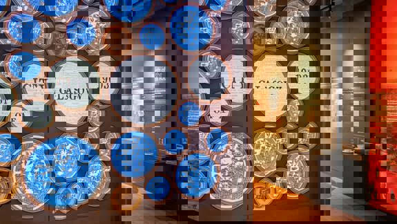 Exhibit display featuring blue and white decorative plates with text reading “City of Glasgow 1903” at the Willow Tea Rooms exhibition. A nearby wall display titled “Birth of the Tea Room” includes historical information and a world map.