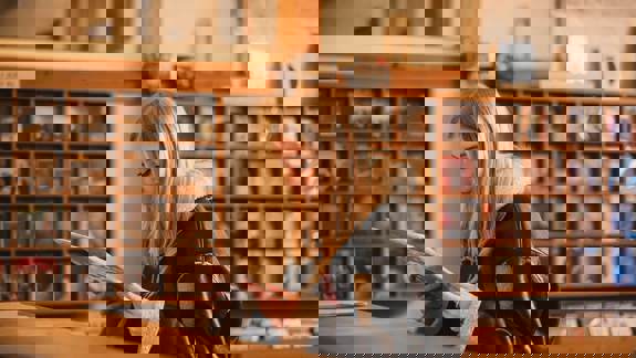 A person with long blond hair looks at a record album, with rows of albums visible on shelves in the background.