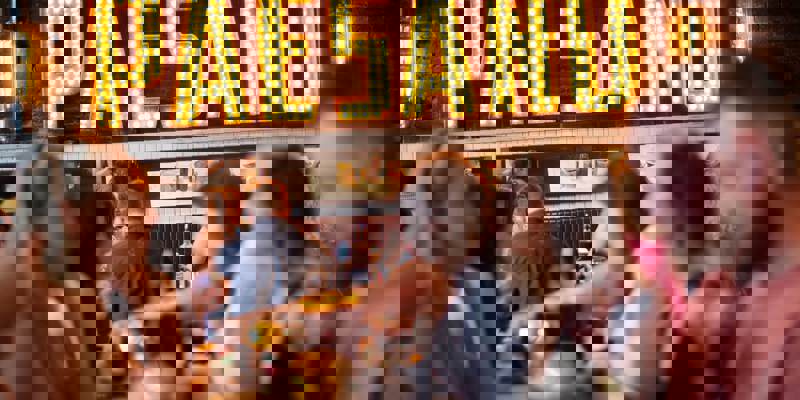 A group of people sitting at tables in a restaurant, with a large, lighted sign that reads "PAESANO" in the background.