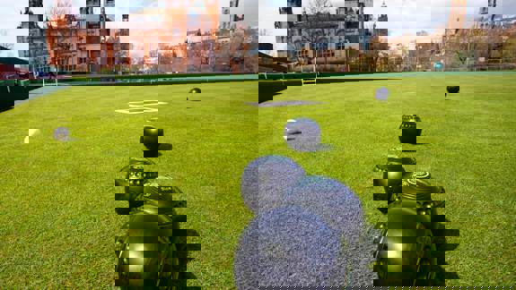 A lawn bowls court with several bowls scattered on the grass. In the background, there is a large, classic building with a red facade and a blue sky with some clouds.