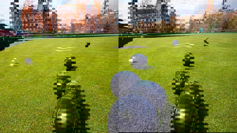 A lawn bowls court with several bowls scattered on the grass. In the background, there is a large, classic building with a red facade and a blue sky with some clouds.