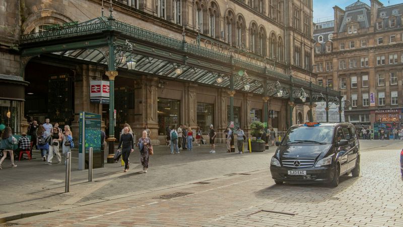 People walking outside a large historic train station with a taxi driving on a cobblestone street.