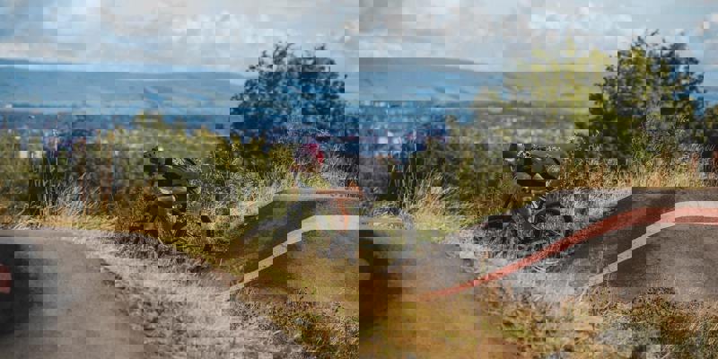 A mountain biker in a black t-shirt and red helmet rides on a dirt track with a city and hills in the background.