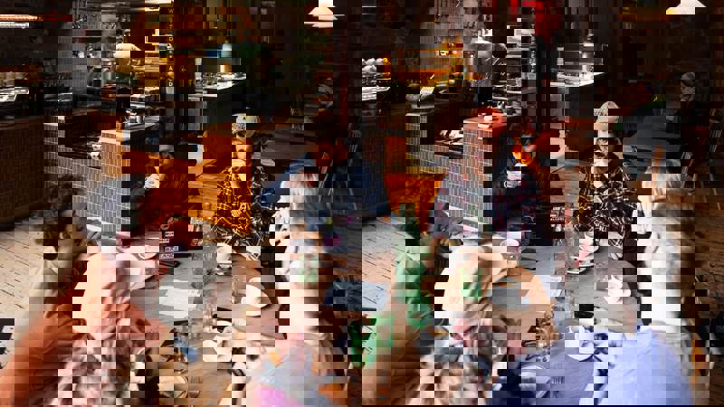 A group of six people sit around a round wooden table in a busy, rustic-industrial Outlier.   They are drinking coffee and talking, with an open kitchen visible in the background.