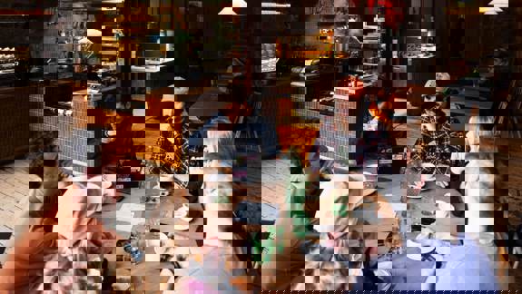 A group of six people sit around a round wooden table in a busy, rustic-industrial Outlier. They are drinking coffee and talking, with an open kitchen visible in the background.