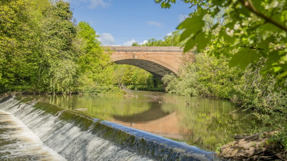 A wide stone bridge crosses a river surrounded by green trees. The river flows into a mini waterfall.