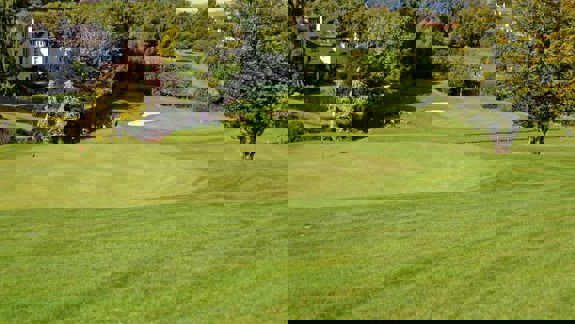 Sunny golf course with smooth greens, surrounding trees, nearby houses, and a few people walking along a path near the fairway.