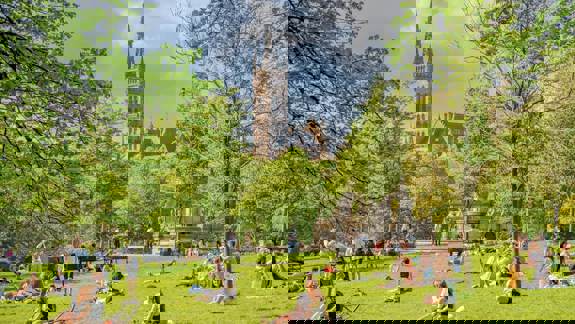 People relax on a sunny day in a green park with a grand, spired building visible in the background.