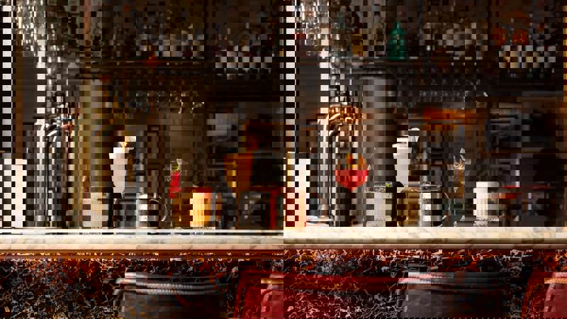 Five distinct cocktails sit on a marble-topped bar with brass fittings, set against a backdrop of neatly stacked bottles and barware.