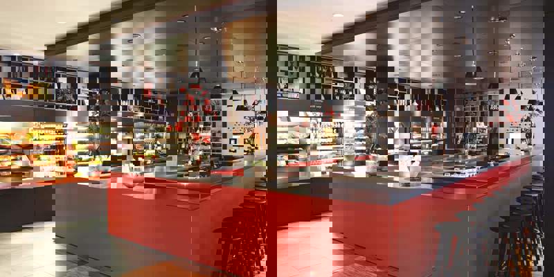 Bright, modern hotel canteen or bar area with a long, vibrant red counter, black bar stools, and shelving displaying food, drinks, and decor.