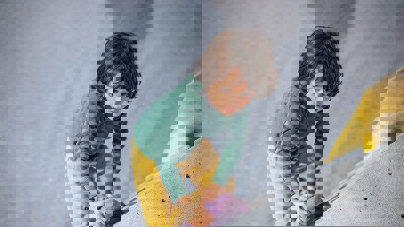 A young child in a green shirt and yellow trousers indoor climbing, gripping a purple handhold on a grey climbing wall.