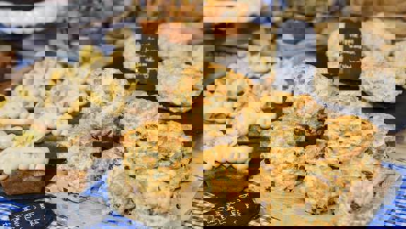 A close-up shows a display of baked goods, including a pile of golden raisin scones on a wooden board, banana  and peanut butter loaves, and vegan apple and cinnamon scones, all artfully arranged for sale.