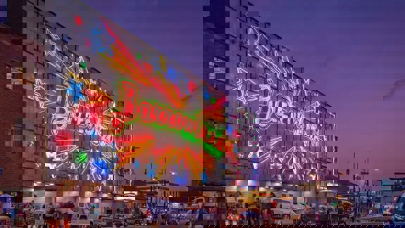 The famous Barrowland sign which says the word Barrowland surrounded by shooting stars is lit up against a dusk sky. People mingle at the front of the venue.