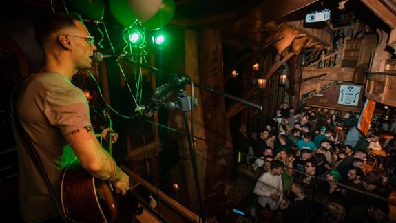 A musician with a guitar and glasses stands on a stage, singing into a microphone. A crowd of people watches the performance onstage from below in a large, wood-panelled pub.
