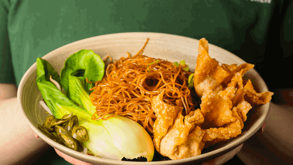Bowl with crispy wontons, stir-fried noodles, bok choy, and sliced green chillis, held by person wearing green “VEGAN KITCHEN” shirt.