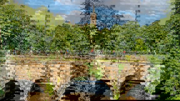 A group of cyclists riding across a stone bridge over a river, with trees and a distant tower visible behind them.