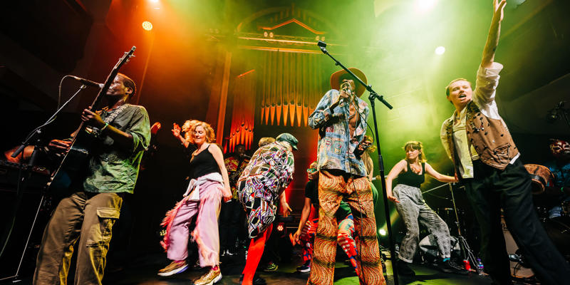 A band and dancers performing on a colourful stage, with the main singer in a hat raising an arm.