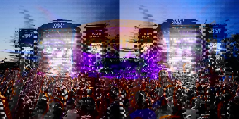 A massive crowd with raised hands faces a brightly lit TRNSMT main stage at twilight, illuminated by vivid purple lights and screen displays.