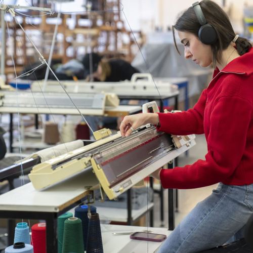 A woman in a red sweater and headphones focuses intently on a knitting machine in a textile workshop. Various colored yarns are visible in the foreground.
