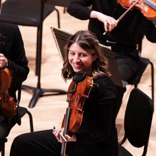 Two violinists in an orchestra seating, smiling warmly and holding violins. The scene is bright, with wooden floors and music stands nearby.