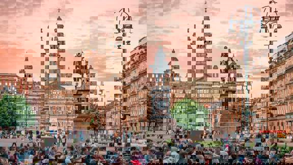 George Square in Glasgow at sunset, with a crowd of people gathered in the foreground and the illuminated City Chambers building and statues in the background.
