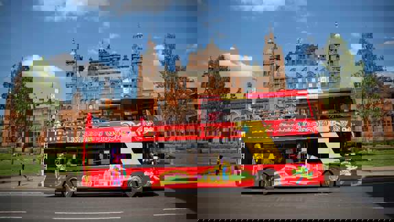 A double-decker bus with "City Sightseeing Glasgow" written on its side, driving past a large, red brick building.