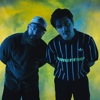 Two men posing against a blue and yellow background, leaning slightly forward toward the camera under studio lighting.