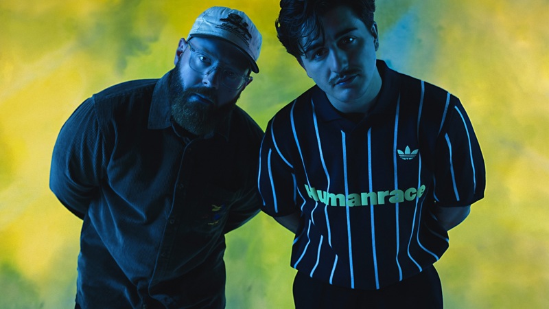Two men posing against a blue and yellow background, leaning slightly forward toward the camera under studio lighting.