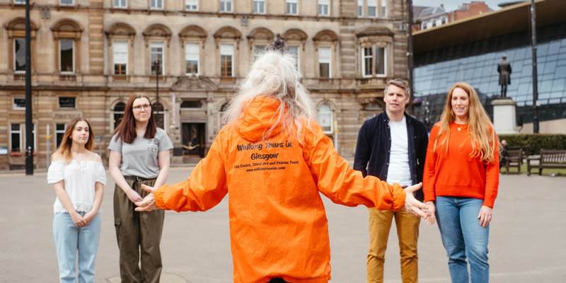 A woman in an orange "Walking Tours in Glasgow" jacket with outstretched arms addresses a small group of people in a city square, with historic buildings in the background.