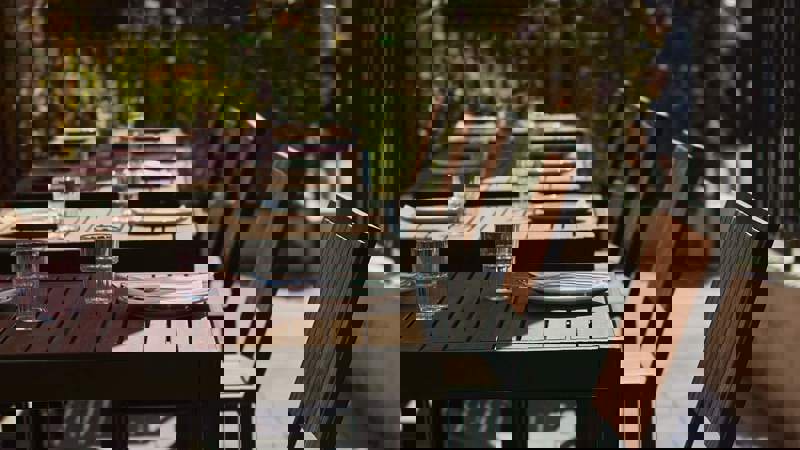 Outdoor dining area with a wooden table set with glasses and plates, surrounded by chairs under a red umbrella.