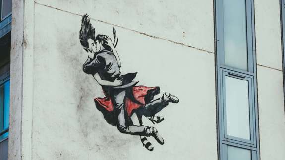 A stencil-style mural on a white building wall by The Rebel Bear, depicting two figures locked in a romantic embrace while appearing to fall or float through the air.