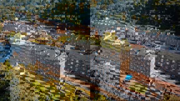 An aerial view of New Lanark, a 19th-century cotton mill village, with large stone buildings surrounded by green trees and a river.