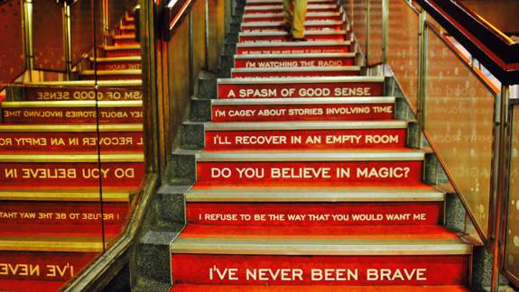 An interior view of red stairs, each riser painted with white song lyrics from the band Biffy Clyro, including "I'll recover in an empty room."