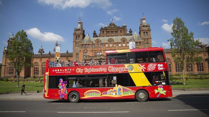 A red, open-top sightseeing bus parked in front of the Kelvingrove Art Gallery and Museum.