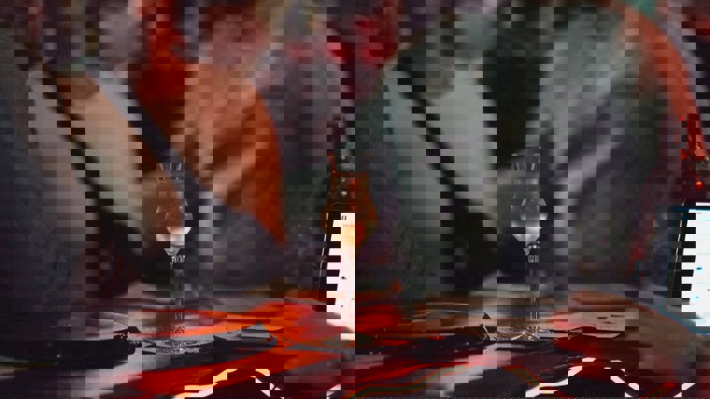 A delicate stem glass holds a pale gold drink on a dimly lit bar, with blurred figures and warm red lighting in the background.