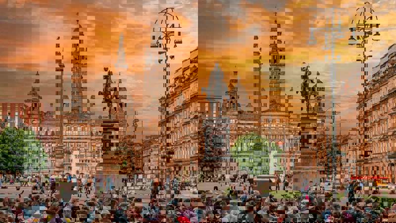 A vibrant photo of a crowd of people gathered in Glasgow's George Square at sunset. In the background, the Glasgow City Chambers stands under a beautiful orange and cloudy sky.