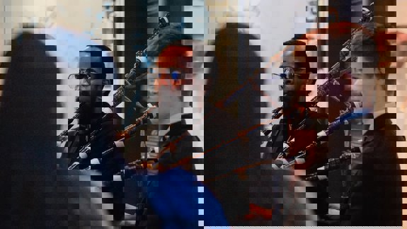 Two people are playing bagpipes indoors. One person has a beard and glasses. The other person is smiling at them and helping them to play the bagpipes. A third person in the foreground with their back to the camera watches the other two..