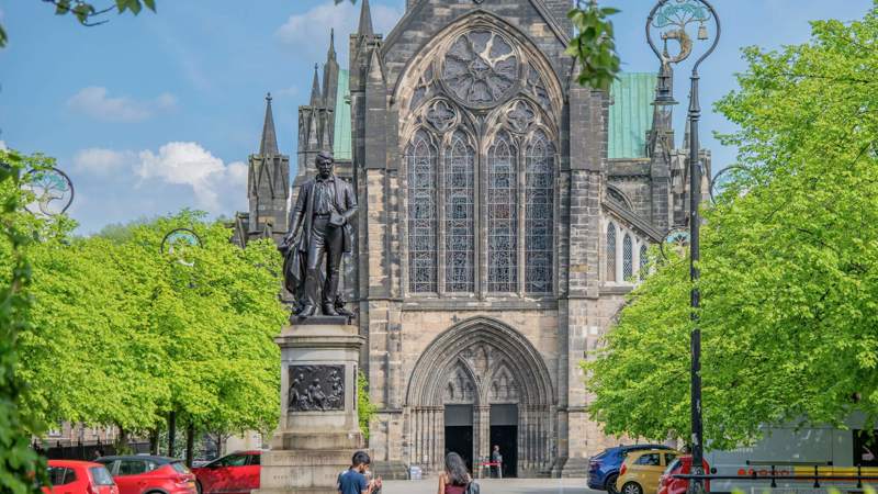 The majestic exterior of Glasgow Cathedral with a large statue in the foreground and green trees and cars around.