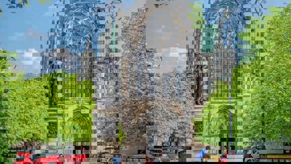 The majestic exterior of Glasgow Cathedral with a large statue in the foreground and green trees and cars around.