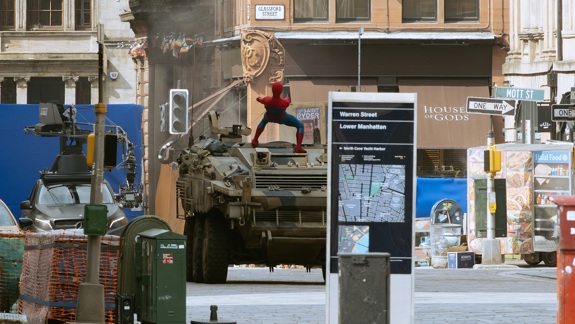 A film set shows a Spider-Man actor on an armoured vehicle in a city street with blue screens, cameras, and crew equipment, simulating a Lower Manhattan scene.