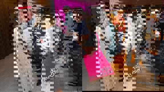 People network at a brightly lit indoor event. A woman in the foreground carries a vibrant pink tote bag prominently featuring the "VISIT GLASGOW" brand in white text.