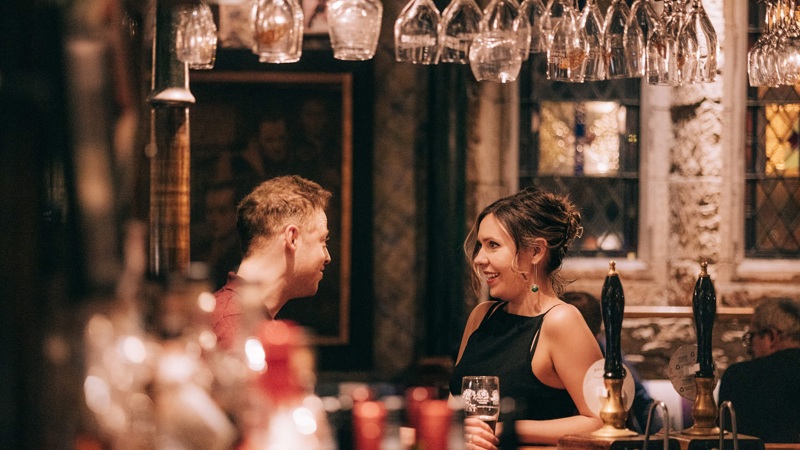 Two people smiling at each other while standing at a pub bar. Lots of upside down wine glasses hang from the bar.
