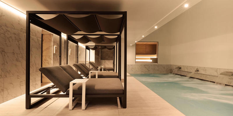 A wide, indoor vitality pool with jets and underwater lighting in a serene, dark-tiled spa setting.