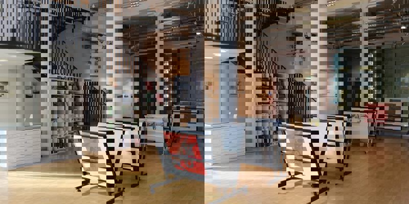 An empty art studio and gallery with a light wooden floor. The space has a staircase, white support columns, and racks displaying framed artwork.