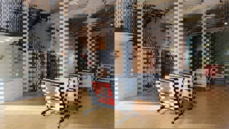 An empty art studio and gallery with a light wooden floor. The space has a staircase, white support columns, and racks displaying framed artwork.