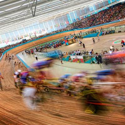 A wide-angle, blurred shot of a cycling race inside a velodrome. Cyclists are racing around a large, wooden track. The stands are filled with spectators.